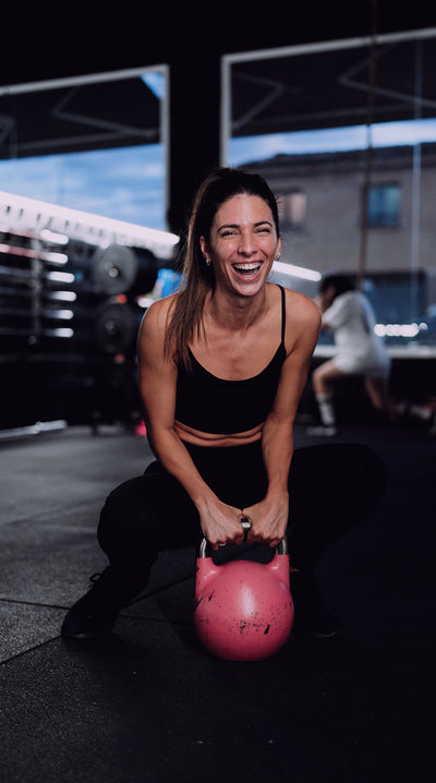 Mujer sonriendo en cuclillas en un box de crossfit con una kettlebell rosa 